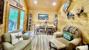 A cozy wooden cabin interior with a dining area, two sofas, and a mounted TV. - Riverfront Cabin Downtown Helen! Hot Tub, FirePit,Arcade,Tubing & Private Park!♥️ (Helen)