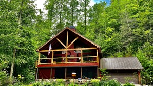 A two-story log cabin with a balcony, surrounded by lush greenery and a small adjacent shed. - Riverfront Cabin Downtown Helen! Hot Tub, FirePit,Arcade,Tubing & Private Park!♥️ (Helen)