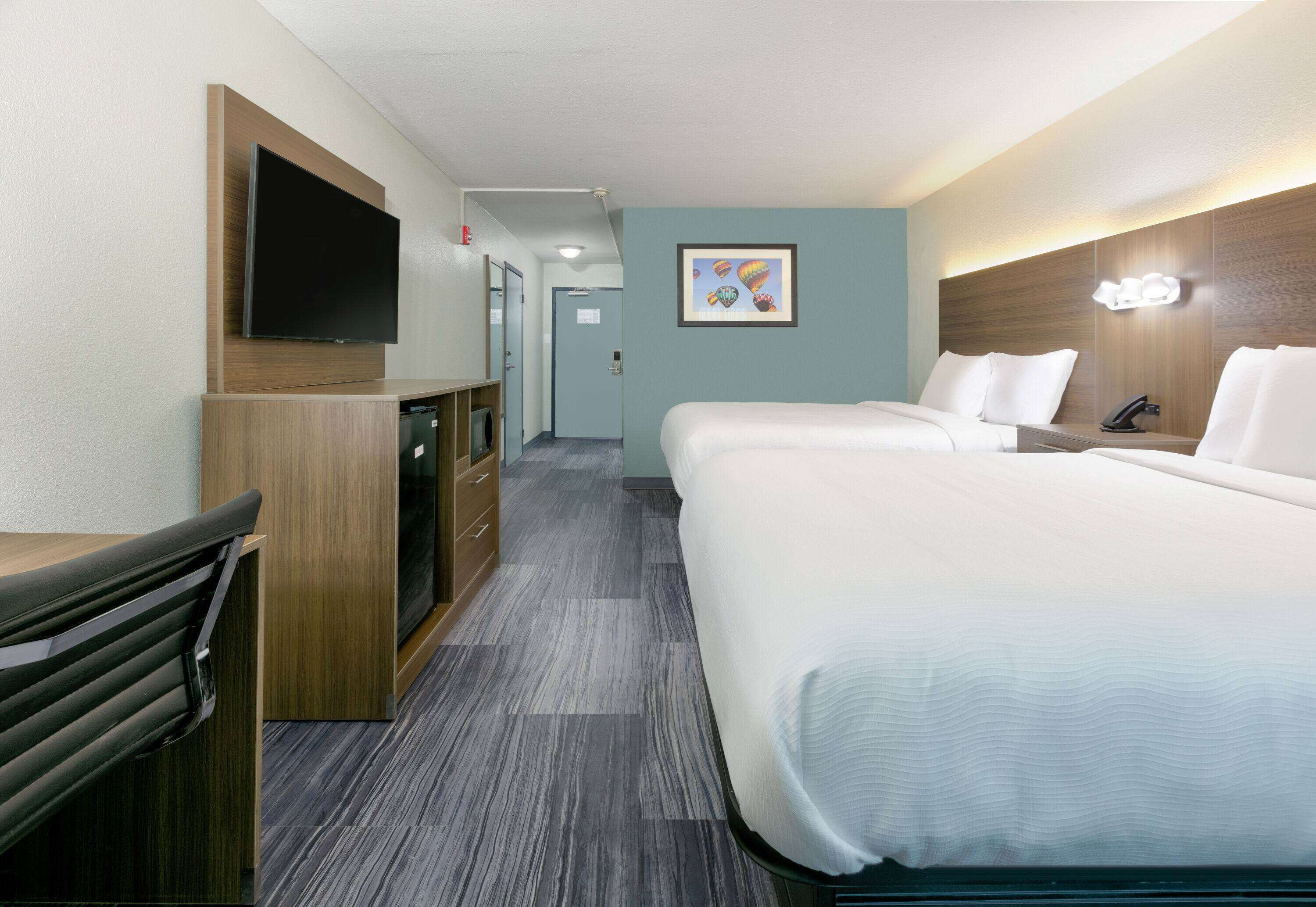 A hotel room with a flat-screen TV mounted on the wall, a desk with drawers, a bed with white bedding, and a framed picture on the wall.
