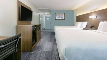 A hotel room with a flat-screen TV mounted on the wall, a desk with drawers, a bed with white bedding, and a framed picture on the wall.