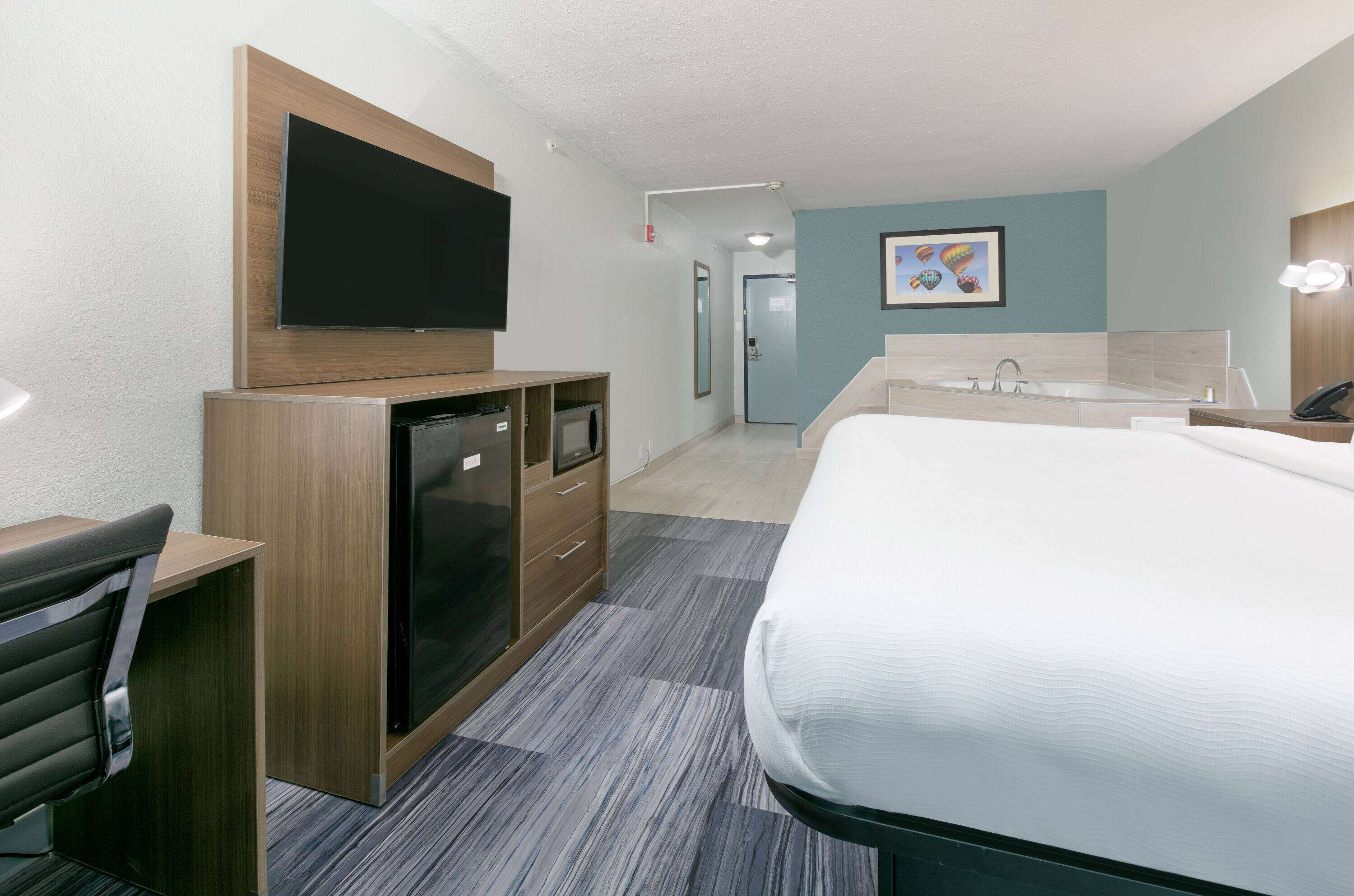 A hotel room with a bed, a desk with a chair, a TV, a minibar, and a bathroom with a sink and mirror.