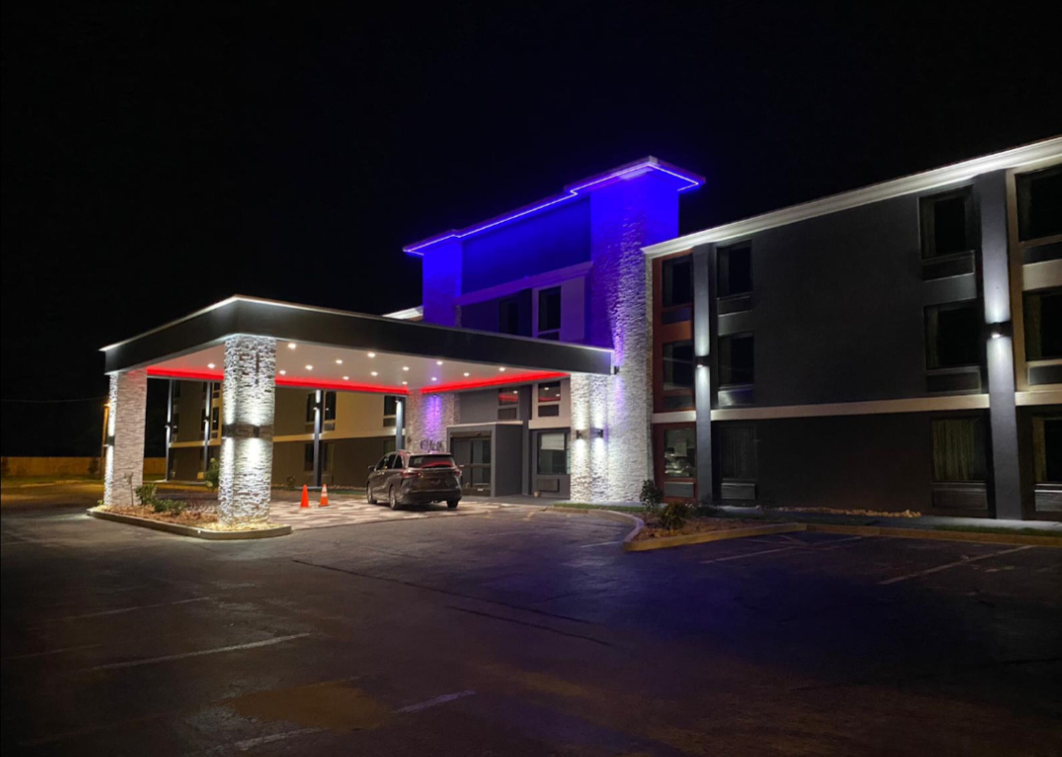 En hotelindgang med et overdækket parkeringsområde, en bil foran og en bygning med blå og røde lys.