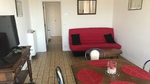A dining area with a wooden table, red placemats, and a glass candle centerpiece. There is a red sofa against the wall and a television on a wooden stand. - Feet in the water  (Langrune-sur-Mer)