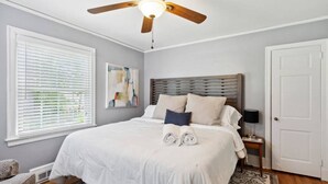 A bedroom with a large bed, a ceiling fan, a window with blinds, a bedside table with a lamp, and a door.