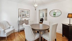 A dining room with a round table, white upholstered chairs, a chandelier, a framed abstract painting, a mirror, and a lamp.