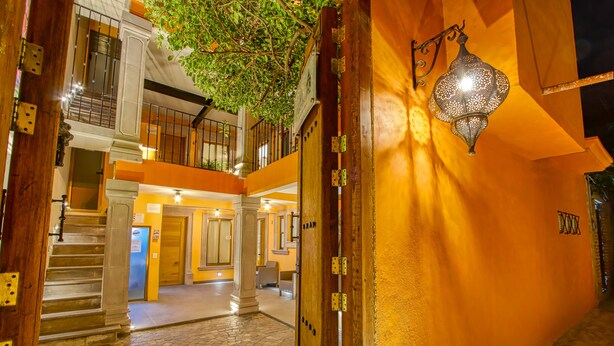 Entrada de hotel con una escalera que conduce al piso superior, una puerta de madera con bisagras metálicas y una lámpara fijada a la pared.