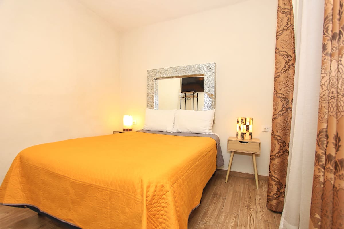 Quarto com cama grande, mesa de cabeceira com abajur, espelho e cortina.