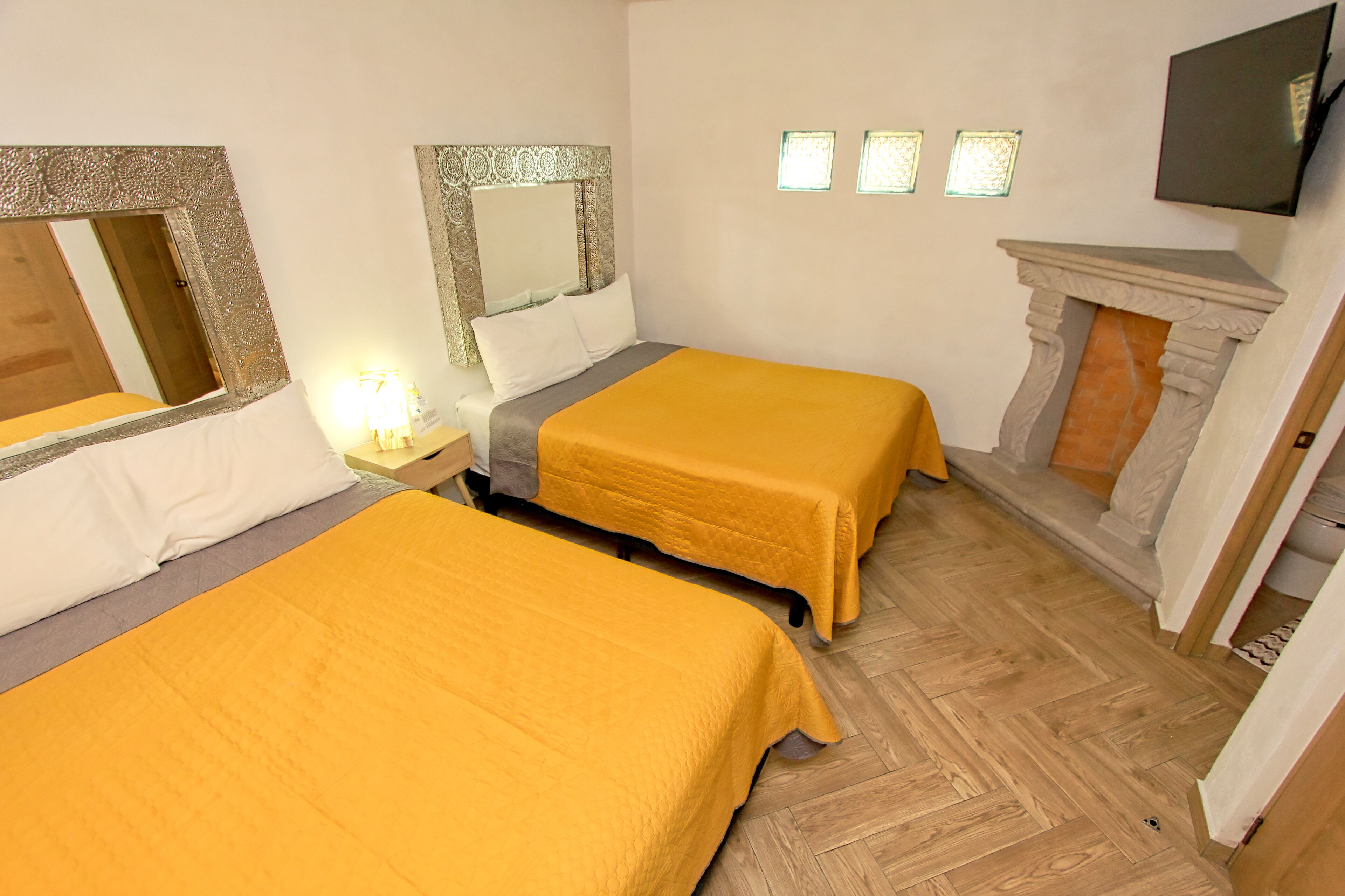 Une chambre d’hôtel avec deux lits, un parquet en bois disposé en point de Hongrie, une cheminée et un téléviseur fixé au mur.