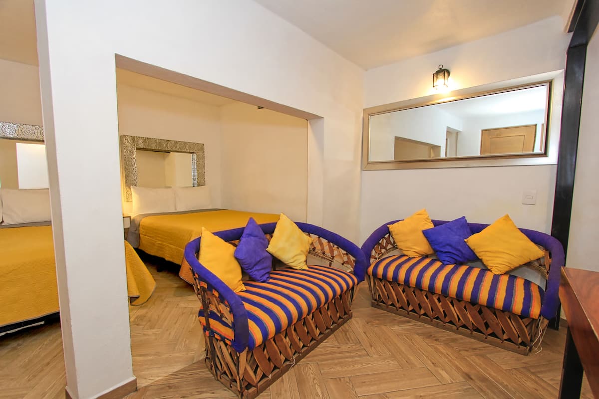 Quarto com dois sofás de vime com almofadas listradas, uma cama com manta amarela e um espelho grande na parede.