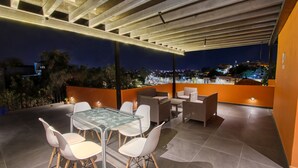 Un patio exterior con una mesa y sillas de vidrio, un sofá y vista a un paisaje urbano de noche.