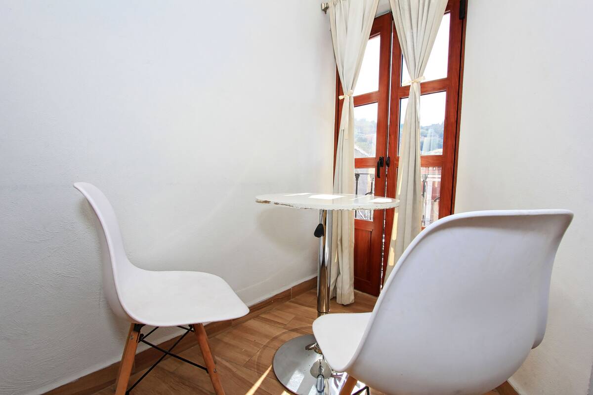A small room with two white chairs and a table, a wooden floor, and a window with curtains.