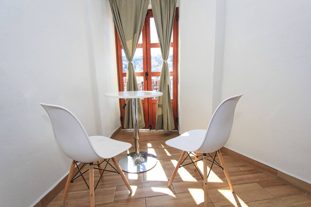 A corner room with two white chairs, a round table, and a view through a window with curtains.