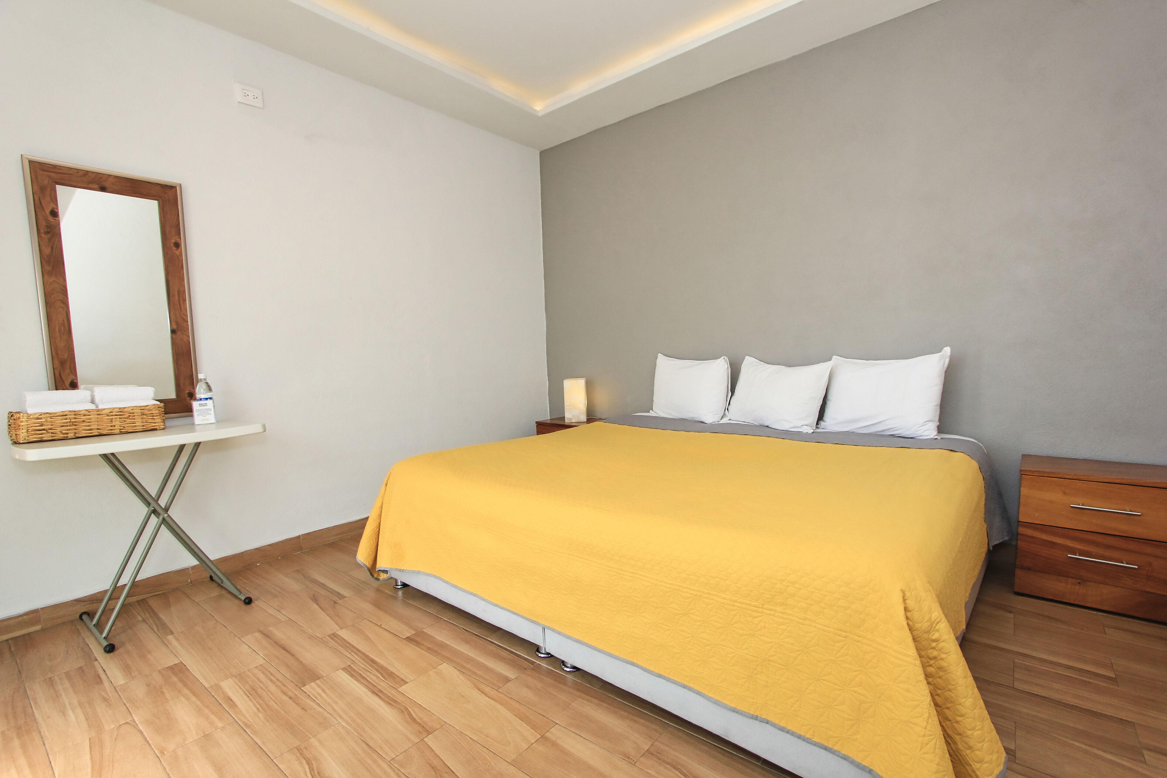Un dormitorio con una cama, una mesita de noche, un espejo y una pequeña mesa con una cesta y una botella.