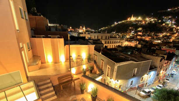 A rooftop terrace with a dining area, potted plants, and a view of a cityscape at night.