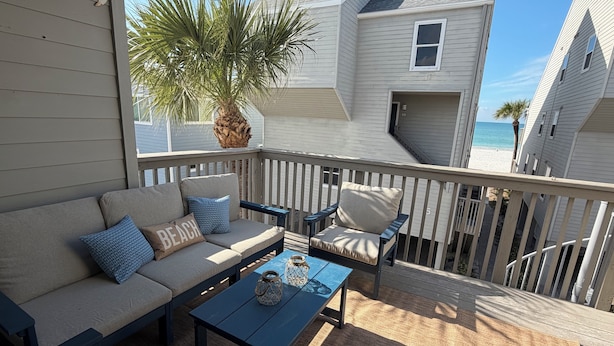 A covered outdoor seating area with a sofa, chairs, and a table, overlooking a beach and neighboring houses. - Pelicans Pointe (Indian Shores)