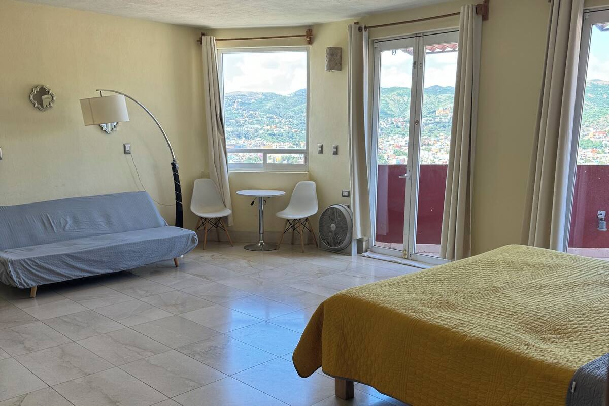 Ein Hotelzimmer mit einem Bett, einem Sofa, einem Tisch und einem Sessel. Ein Fenster bietet Blick auf die Stadt.