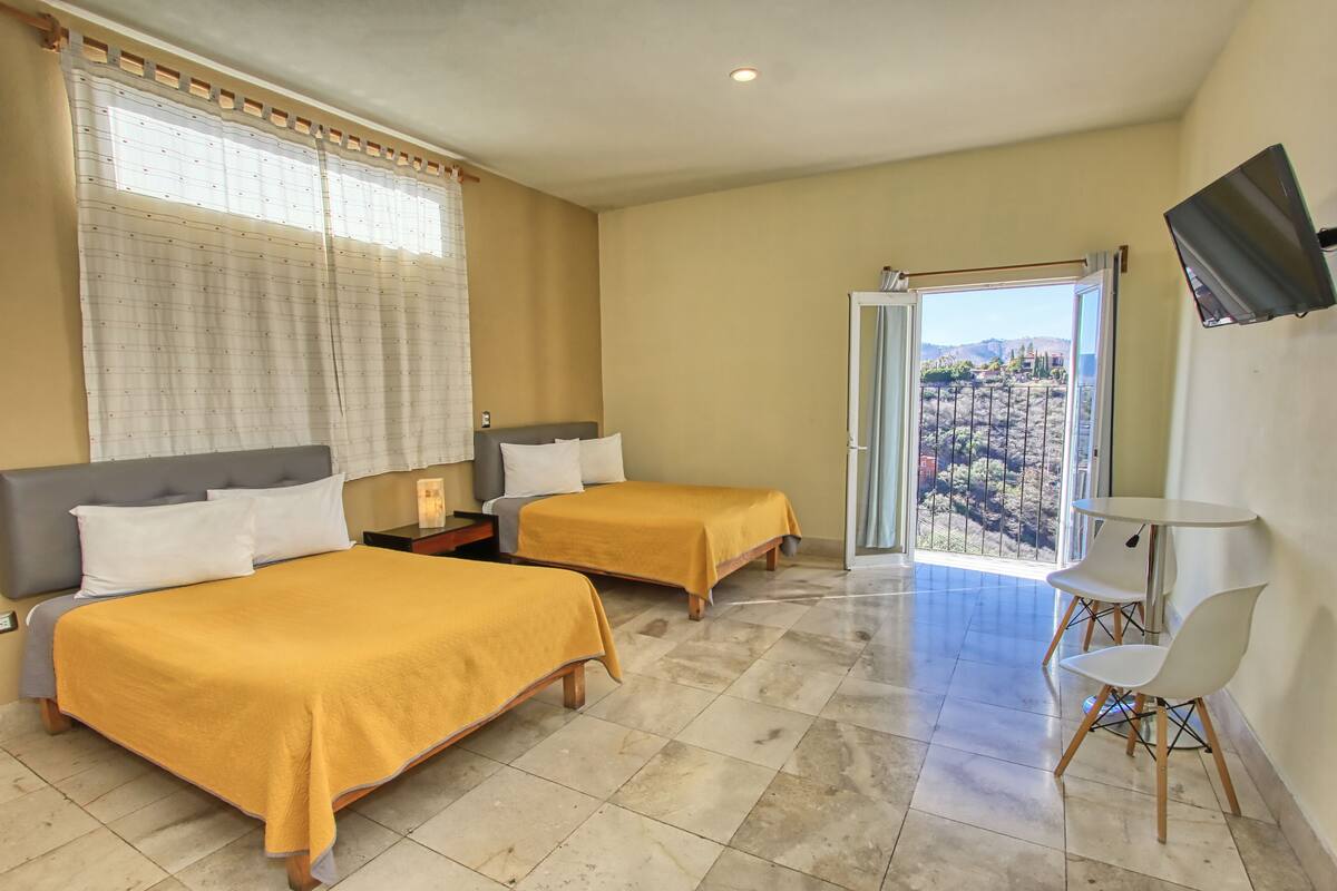 Ein Hotelzimmer mit zwei Betten, einem Fernseher, einem Balkon mit Blick und einem Tisch mit Stühlen.