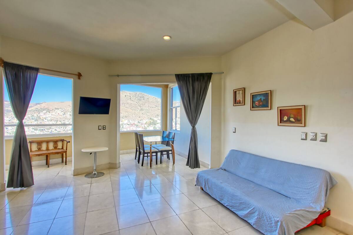 Ein geräumiges Zimmer mit einer Couch, einem Esstisch mit Stühlen, einem Fernseher und einem Balkon mit Vorhängen.