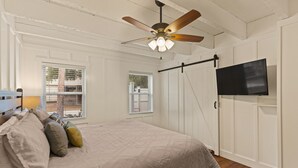 Une chambre à coucher avec un lit, des oreillers, un ventilateur de plafond, un téléviseur fixé au mur et des fenêtres.