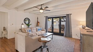 Un salon avec un canapé beige, une petite table ronde, un ventilateur de plafond et une vue sur l’extérieur, visible à travers une porte vitrée.