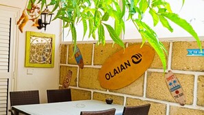 Ein Essbereich mit Tisch, Stühlen, einer hängenden Pflanze, einer Wanduhr und einem Schild mit der Aufschrift 'OLAIAN'.