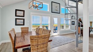 A dining area with a wooden table and wicker chairs, large windows, and a view of the beach.