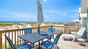 A wooden deck with a table and chairs, overlooking a beach with sand dunes and the ocean.