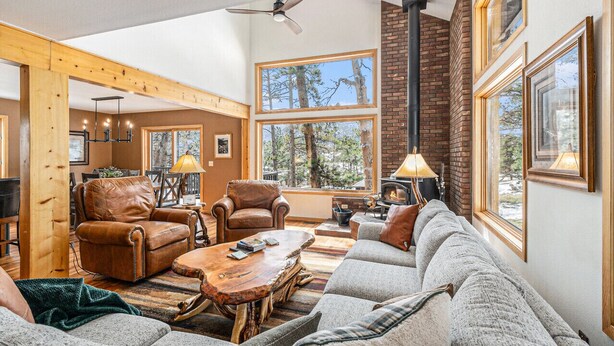 A cozy living room with a fireplace, leather chairs, a sofa, and large windows offering a view of trees. - Luxe Home with Hot Tub & Mountain Views (Estes Park)