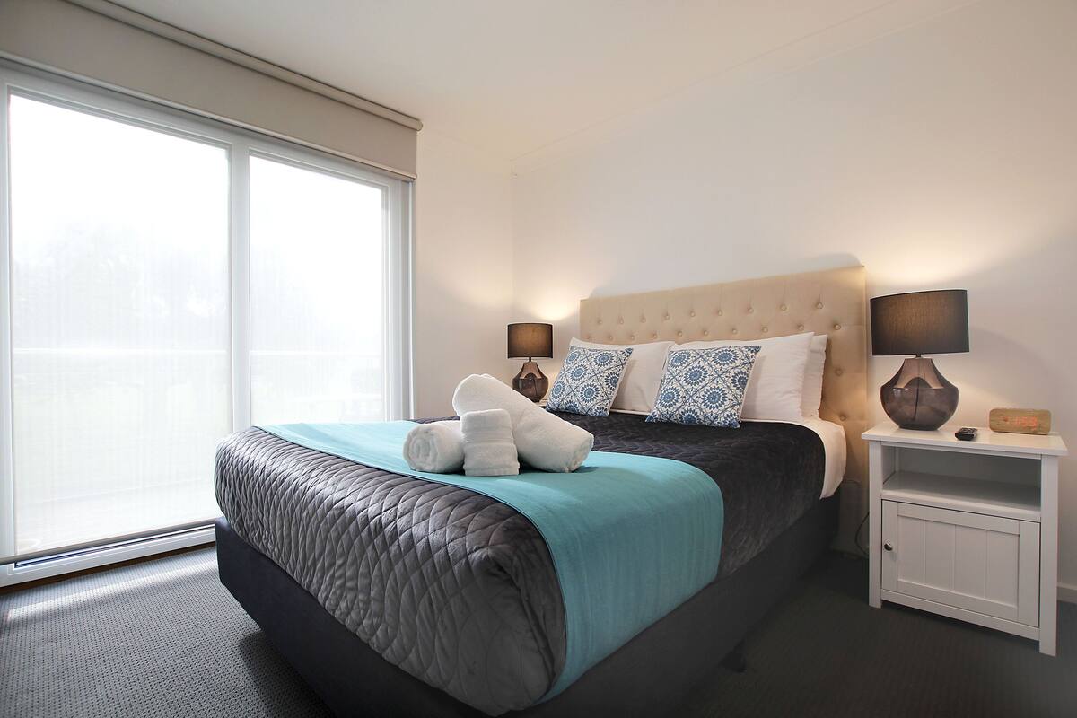 A hotel room with a large bed, two bedside tables, a nightstand, and a window with blinds.