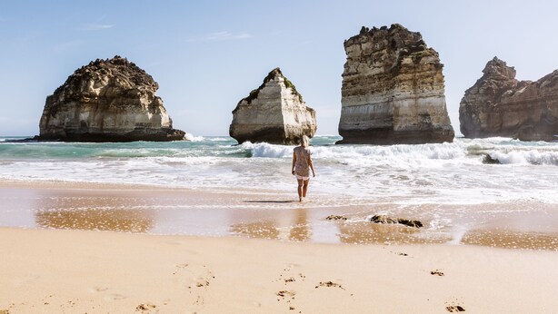 A beach with large rock formations in the background and a person walking on the sand. - Apartments 521 (Warrnambool)