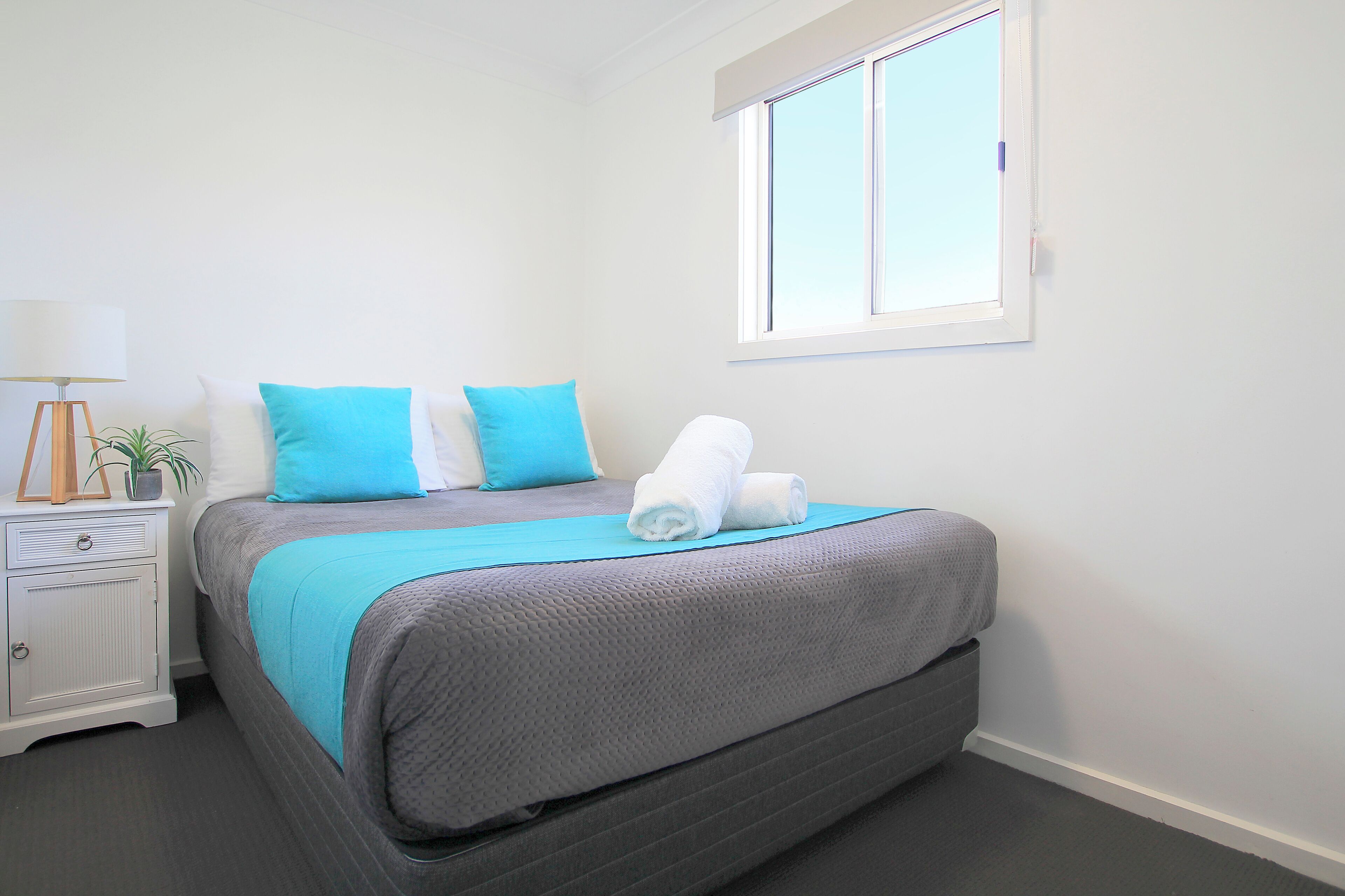 Una cama bien tendida con una colcha gris y azul, una mesita de noche blanca con una lámpara, y una ventana con persianas.