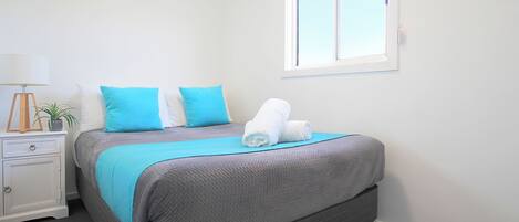 Una cama bien tendida con una colcha gris y azul, una mesita de noche blanca con una lámpara, y una ventana con persianas.