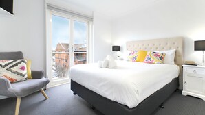 A modern hotel room with a large bed, a grey armchair, a bedside table with a lamp, and a view of the city through a large window. - Apartments 521 (Warrnambool)