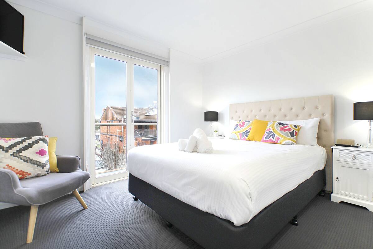 A modern hotel room with a large bed, a grey armchair, a bedside table with a lamp, and a view of the city through a large window.