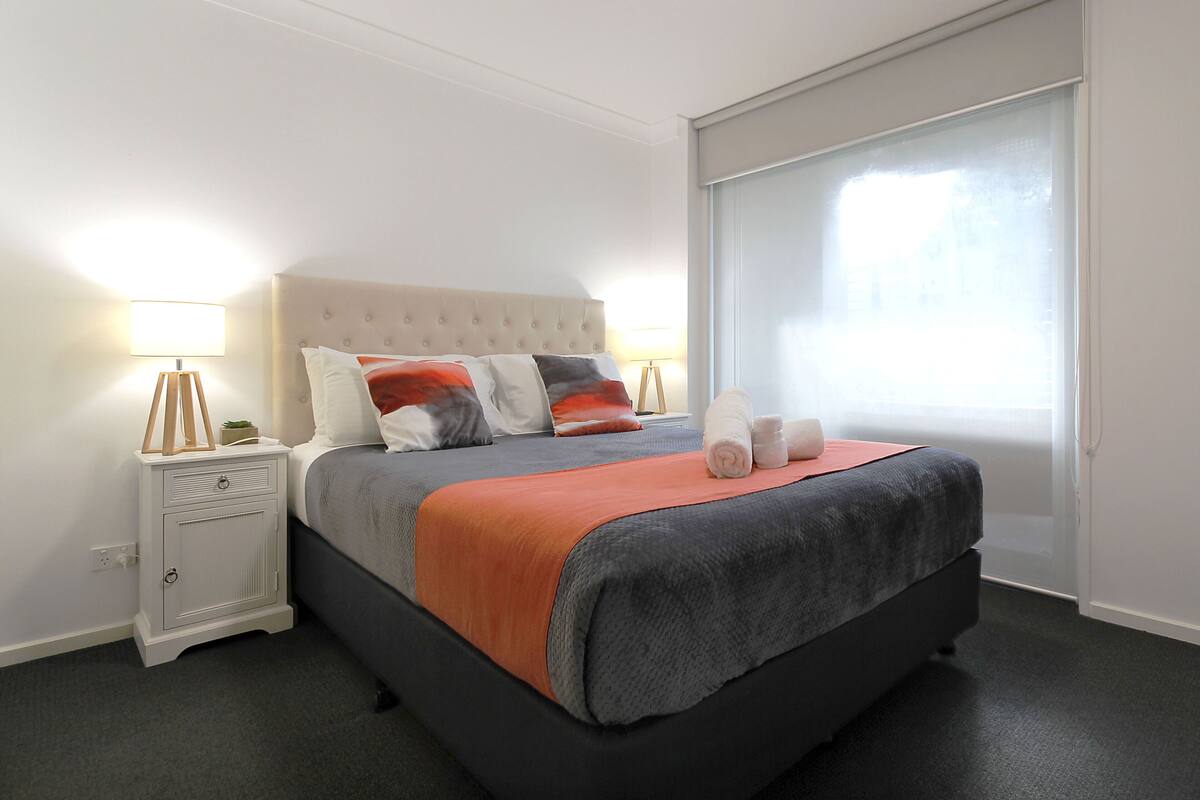 A neatly made bed with a grey and orange blanket, a white bedside table with a lamp, and a large window with blinds.