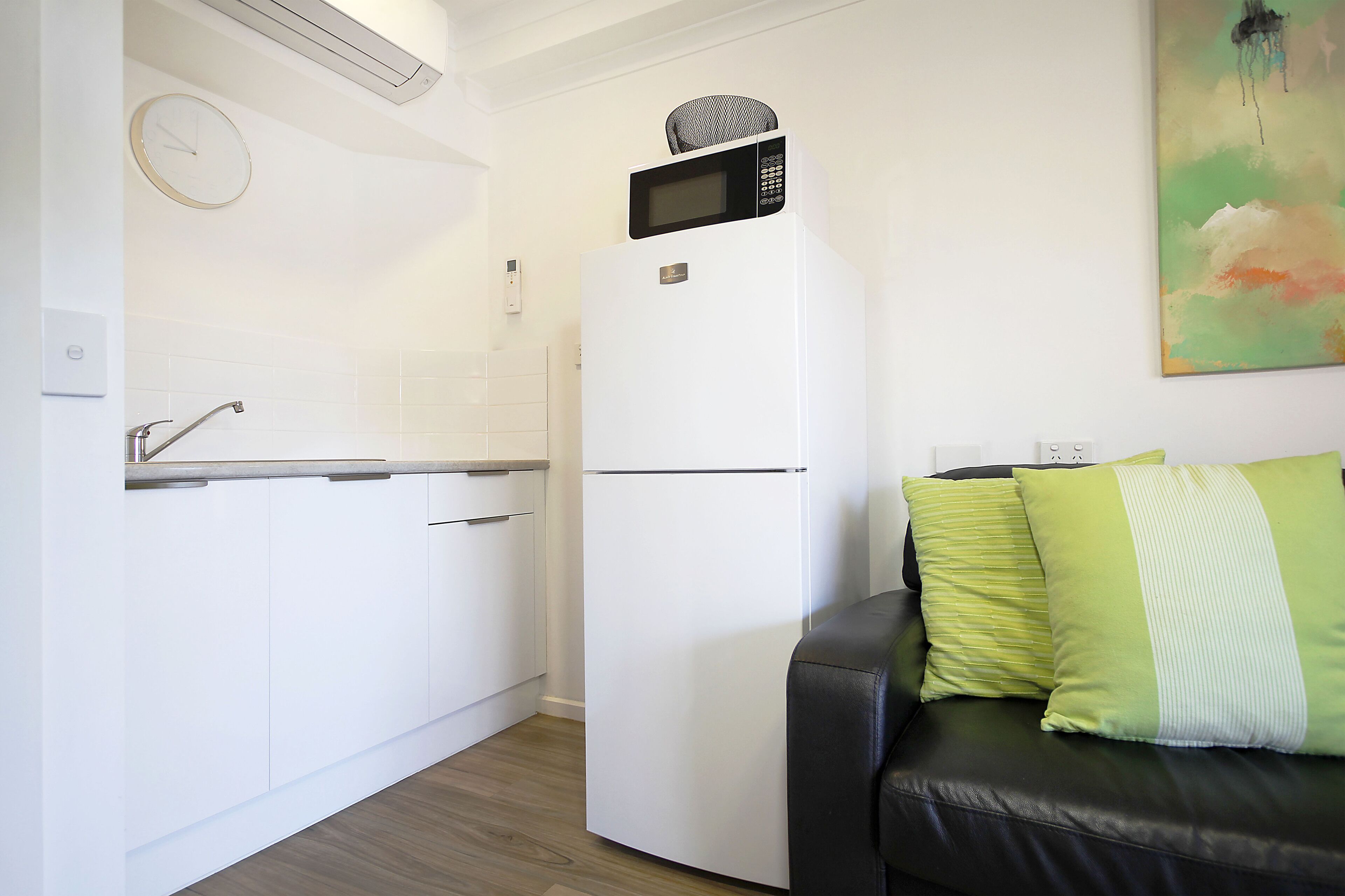 Una cocina compacta con un refrigerador blanco, un microondas encima, un reloj en la pared y un cojín verde y blanco sobre un sofá negro.