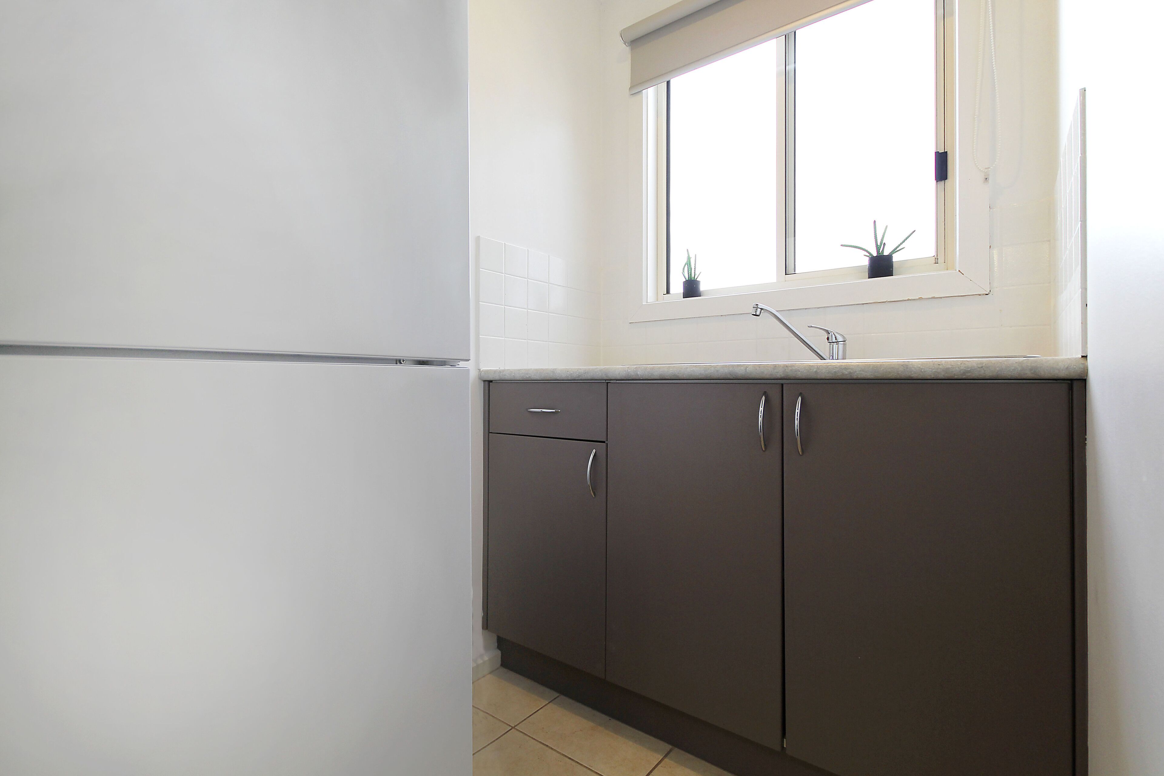 A kitchen with a white refrigerator, dark cabinets, a window with a plant, and a sink.