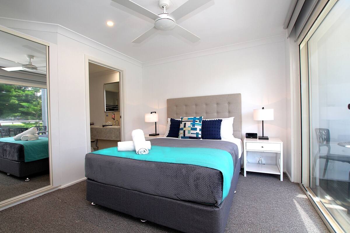 A hotel room with a large bed, a ceiling fan, a nightstand, and a glass door leading to a balcony.