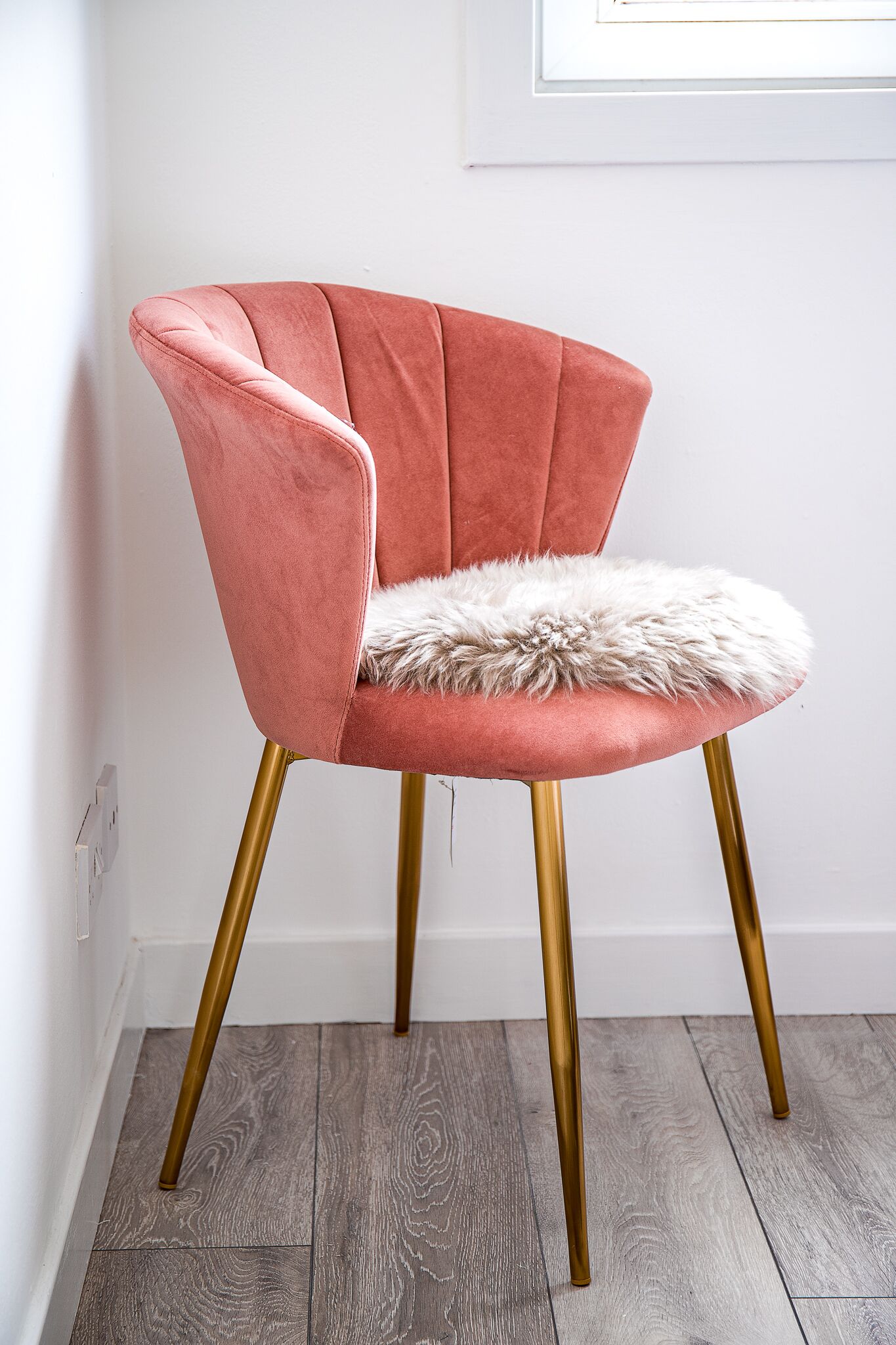 Una butaca rosa tapizada con un asiento mullido y patas doradas, ubicada frente a una pared blanca y suelo de madera.
