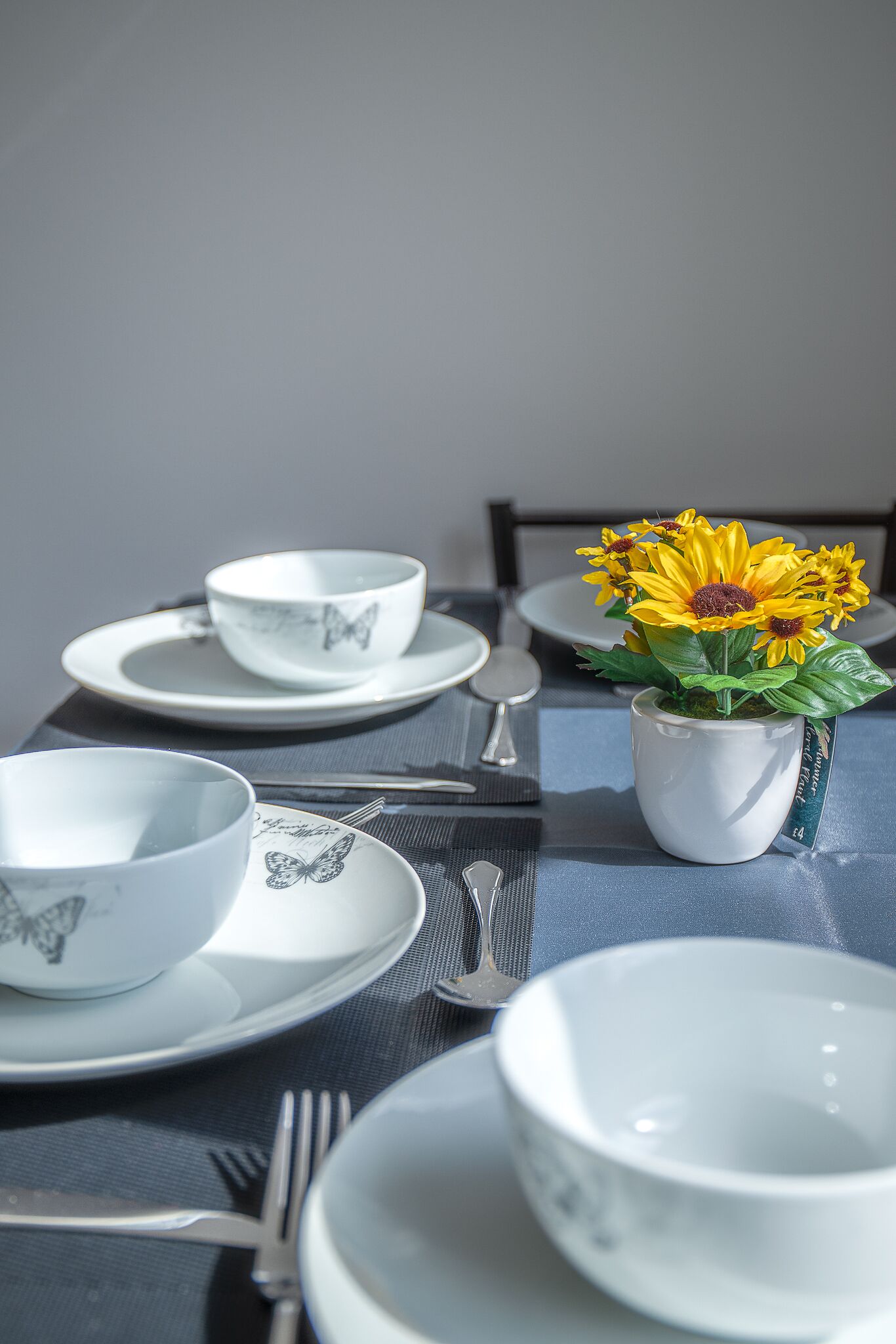 A dining table set with white plates, bowls, and cutlery, with a potted plant of yellow flowers in the center.