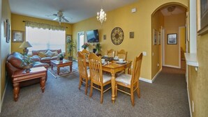 A living room with a sofa, coffee table, and dining table with chairs. There is a ceiling fan, a chandelier, and a TV mounted on the wall. - Windsor_palms_105_sp (Kissimmee)