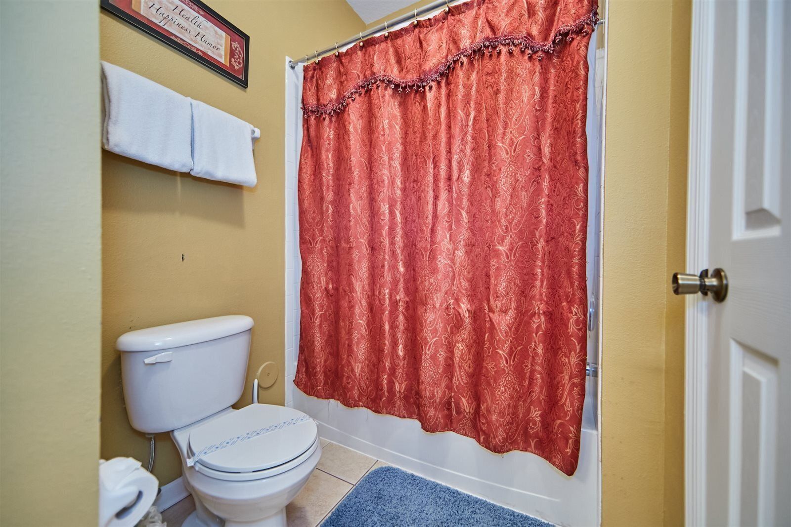 A bathroom with a white toilet, a blue bath mat, and a red shower curtain.