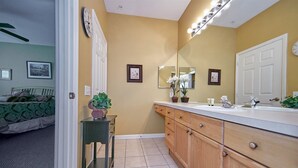 A bathroom with a vanity, sink, and mirror. There is a potted plant on the vanity and a small table with a plant. The walls are yellow, and there is a clock on the wall. - Windsor_palms_105_sp (Kissimmee)