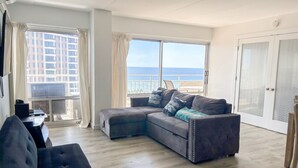 A modern living room with a grey sectional sofa, a small table, and a view of the sea through large windows. - Oceanfront 2BR Condo Ilikai 1822 – Steps to Waikiki Beach | Sleeps 6 | Stunning Views (Waikiki)