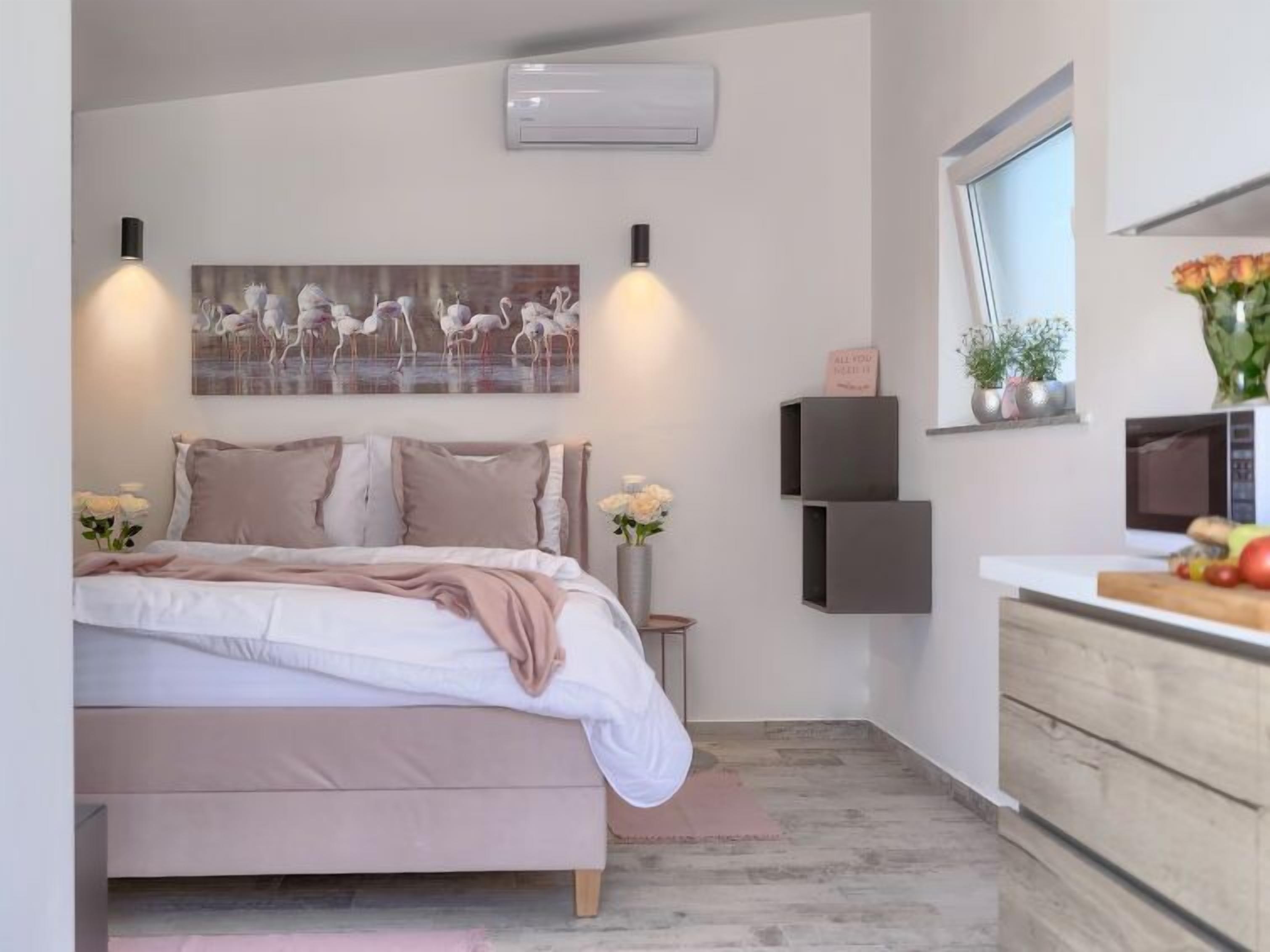 A modern bedroom with a bed, bedside tables, a wall-mounted air conditioner, a painting, and a small potted plant.