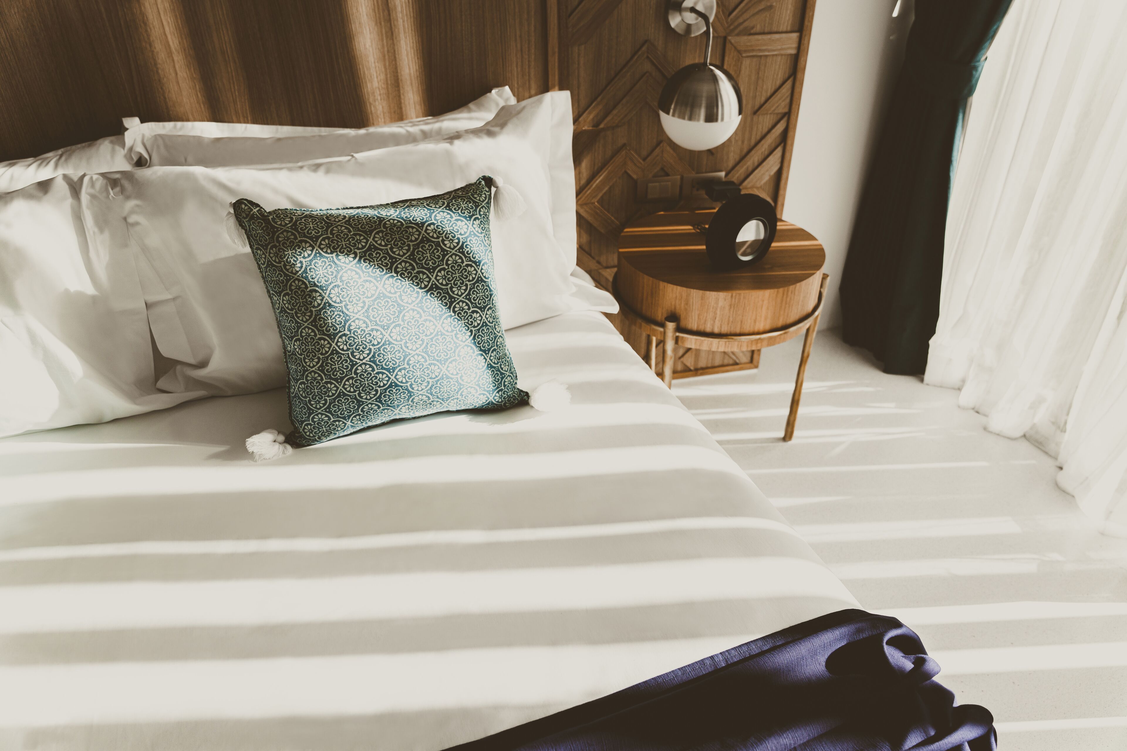 A neatly made bed with white and patterned pillows, a wooden nightstand with a lamp, and a chair.