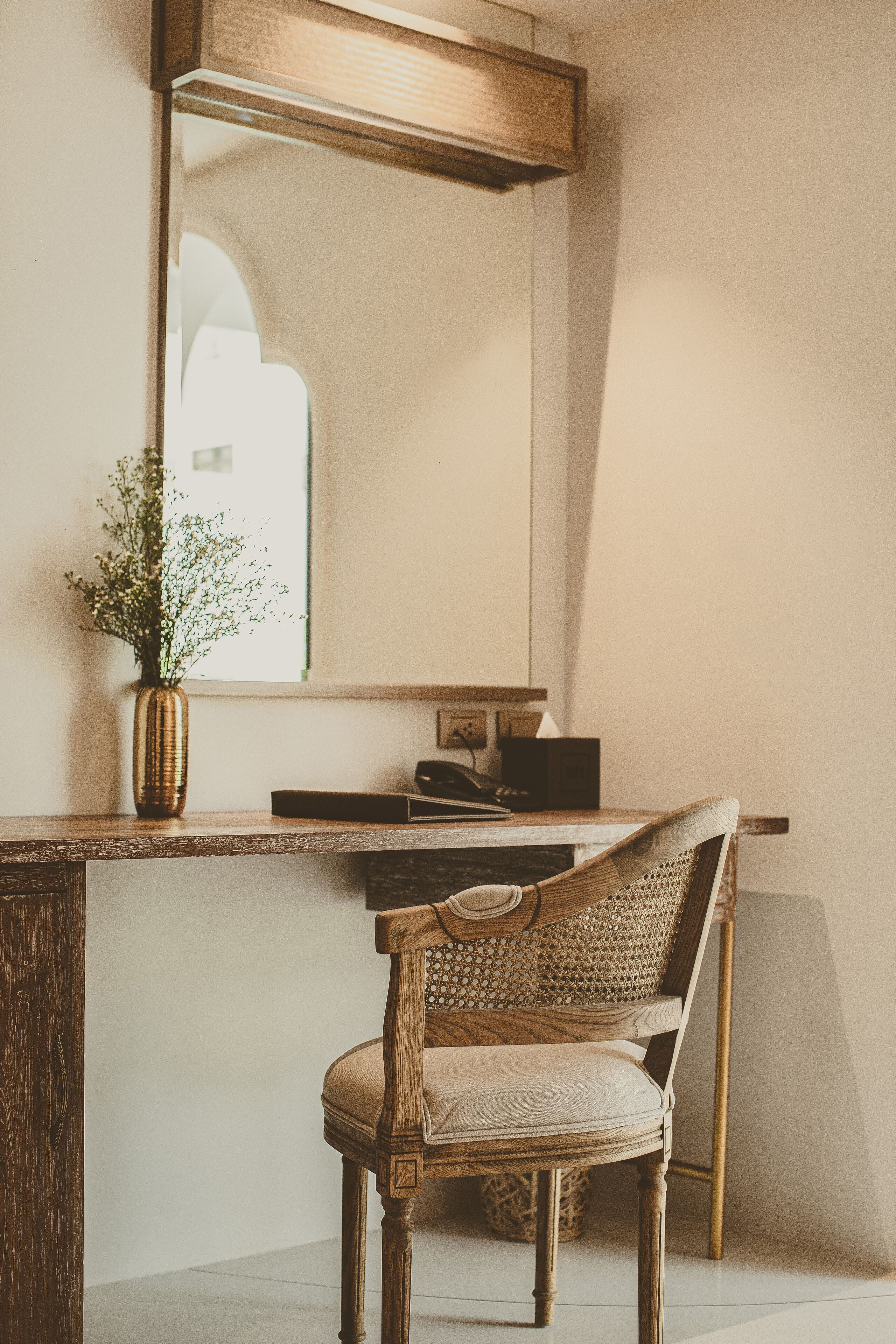 A wooden desk with a vase of flowers, a chair, and a mirror reflecting a window.