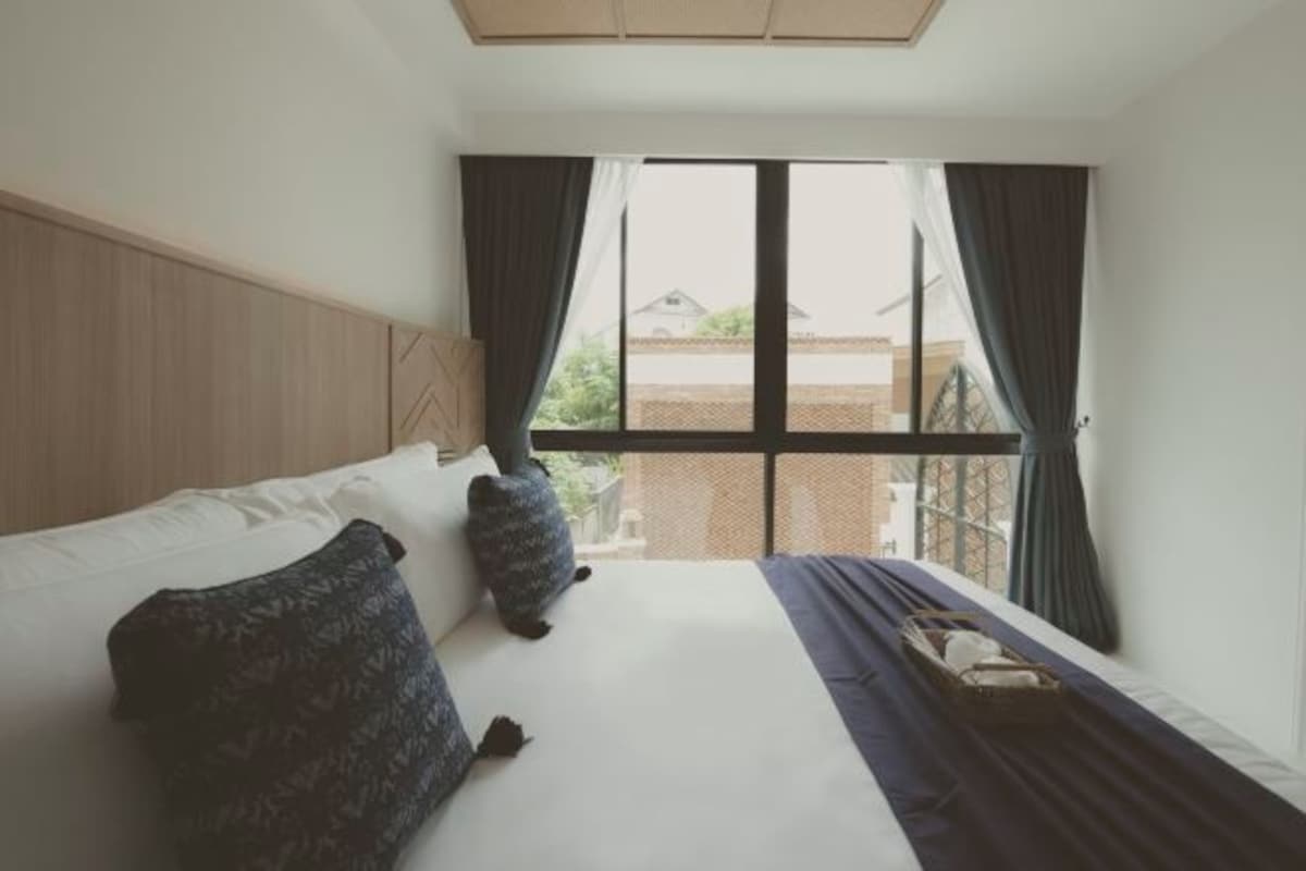 A hotel room with a large bed, two pillows, a window with curtains, and a view of buildings outside.