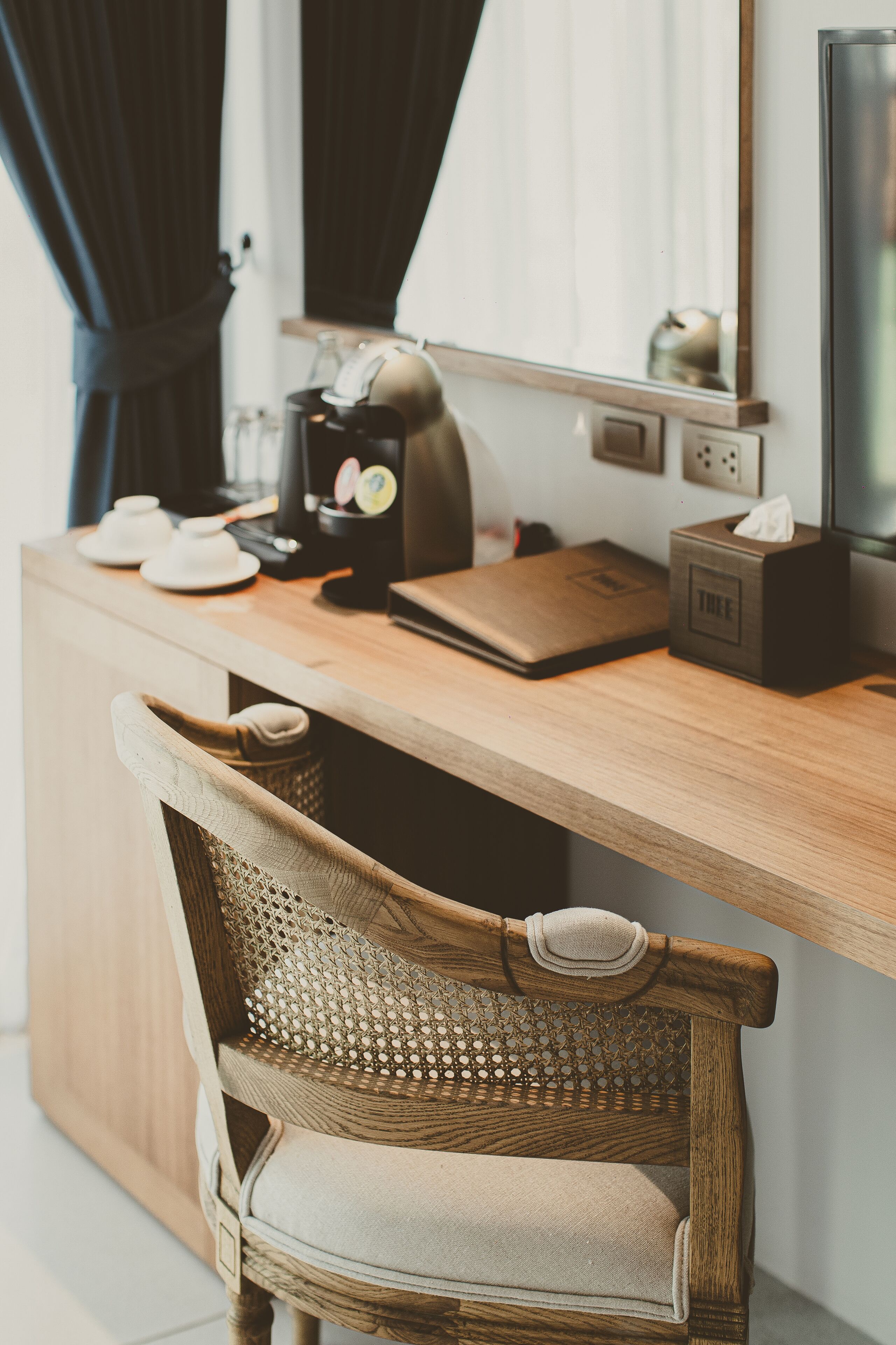 A wooden desk with a laptop, a coffee maker, and a tissue box. A wicker chair is positioned in front of the desk.