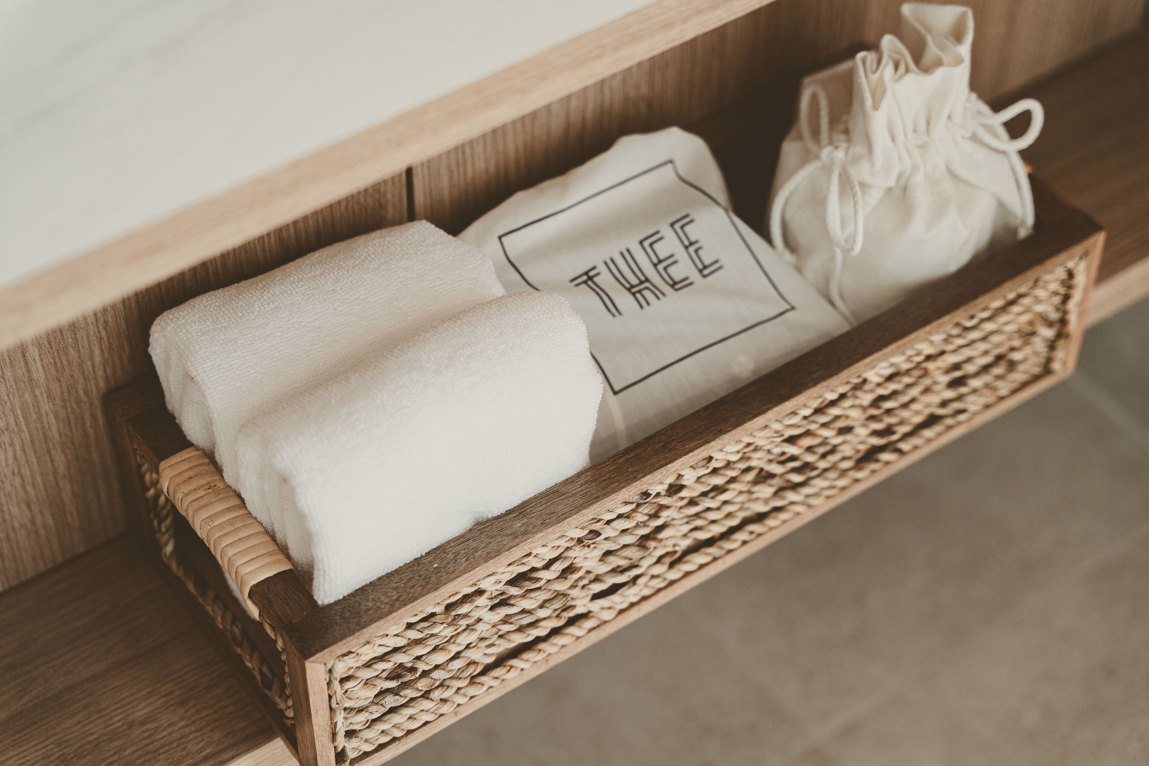 A wooden shelf with a woven basket containing a folded towel and a drawstring bag with the word "TWEE" on it.